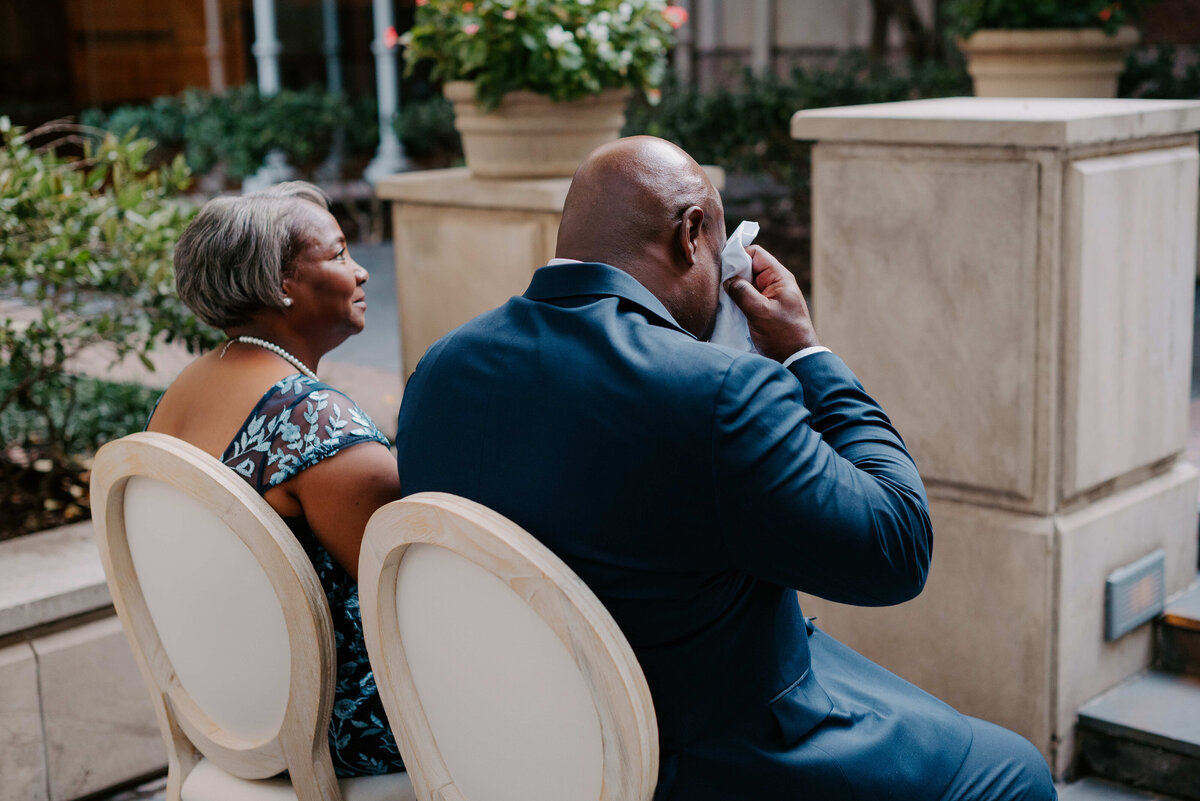 A couple sits outside on white chairs; the man wipes tears with a tissue while the woman smiles gently. The scene is calm and poignant, surrounded by plants.