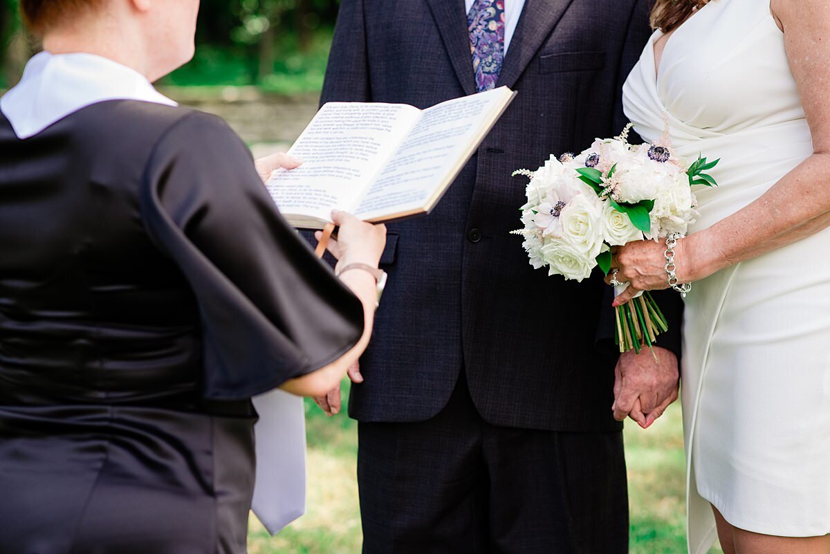 The wedding officiant is wearing a black dress and a white officiant collar as she holds out her book reading the ceremony. The bride and groom look on. The bride is holding a large white floral bouquet and is wearing a short, fitted white dress. The groom is wearing a navy suit.