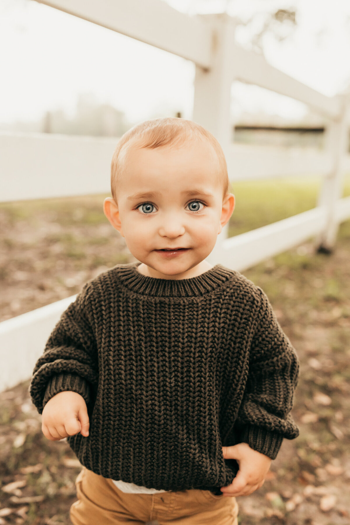 stylish little boy in olive sweater stands against white fence.