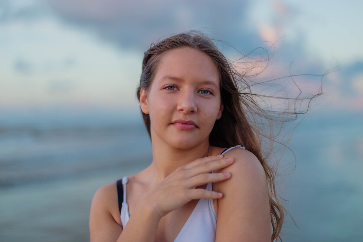 High school senior portrait on the beach at sunrise while the wind blows through her hair gently with her hand  on her shoulder