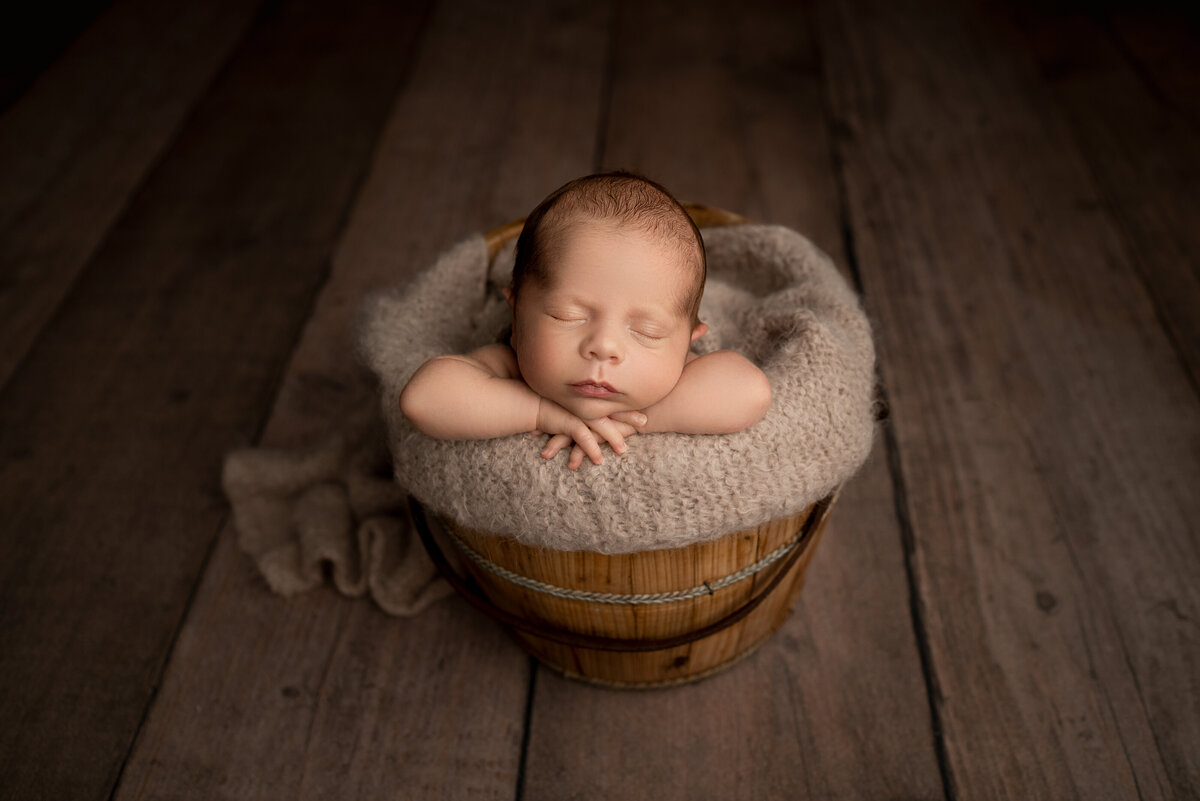 Fine art newborn photos captured by best Main Line newborn photographer Katie Marshall. Baby boy is sleeping in a wooden bucket topped with beige knit fabric. Baby's arms are folded atop the bucket and baby is resting his head on his hands.