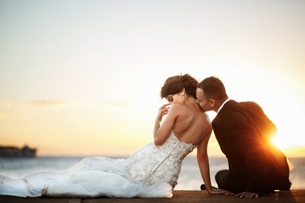 golden-sun-shines-groom-kissing-bride-s-shoulder-while-they-rest-shore