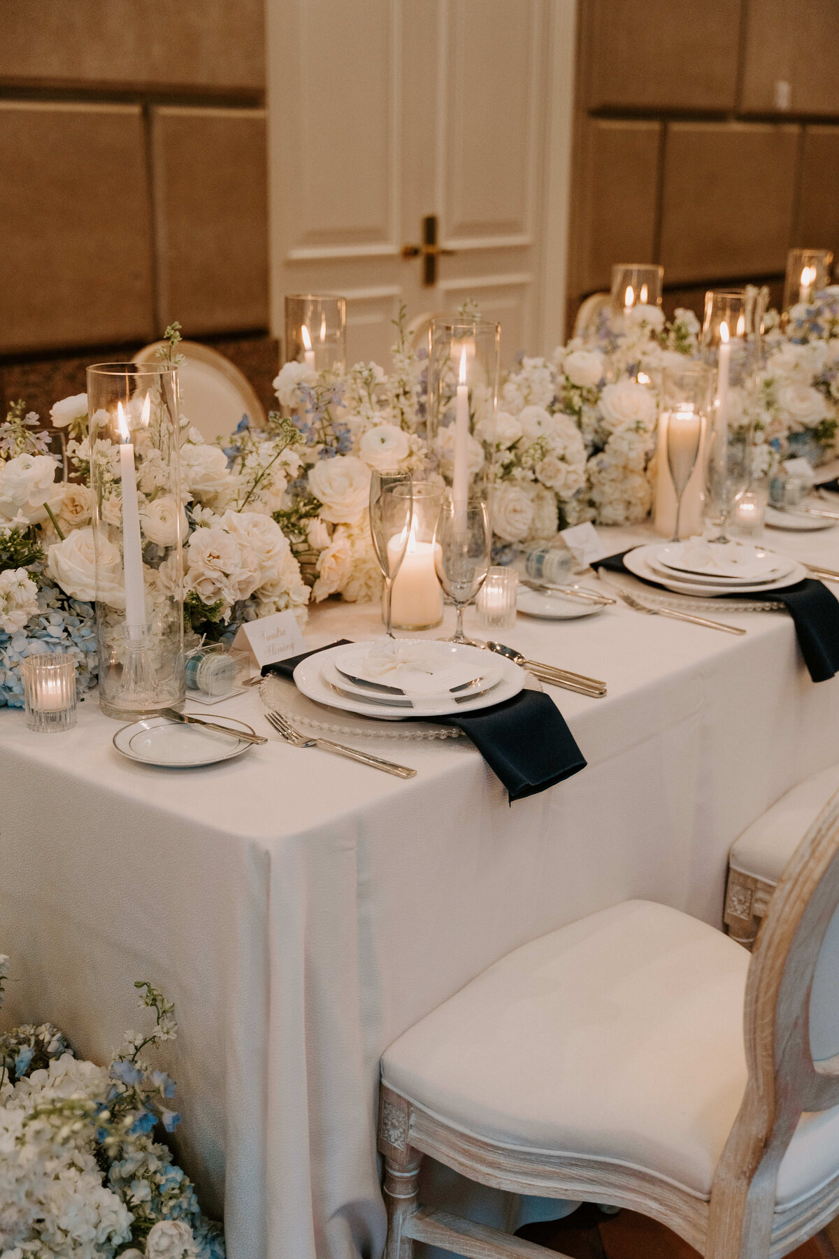 Elegant dining table set with white linens, tall candles, and delicate flower arrangements featuring white roses and greenery, creating a romantic ambiance.