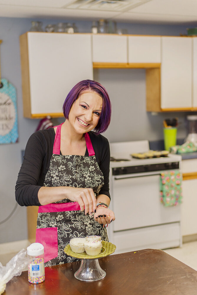 A woman with purple hair and a floral apron smiles while decorating cupcakes in a kitchen.