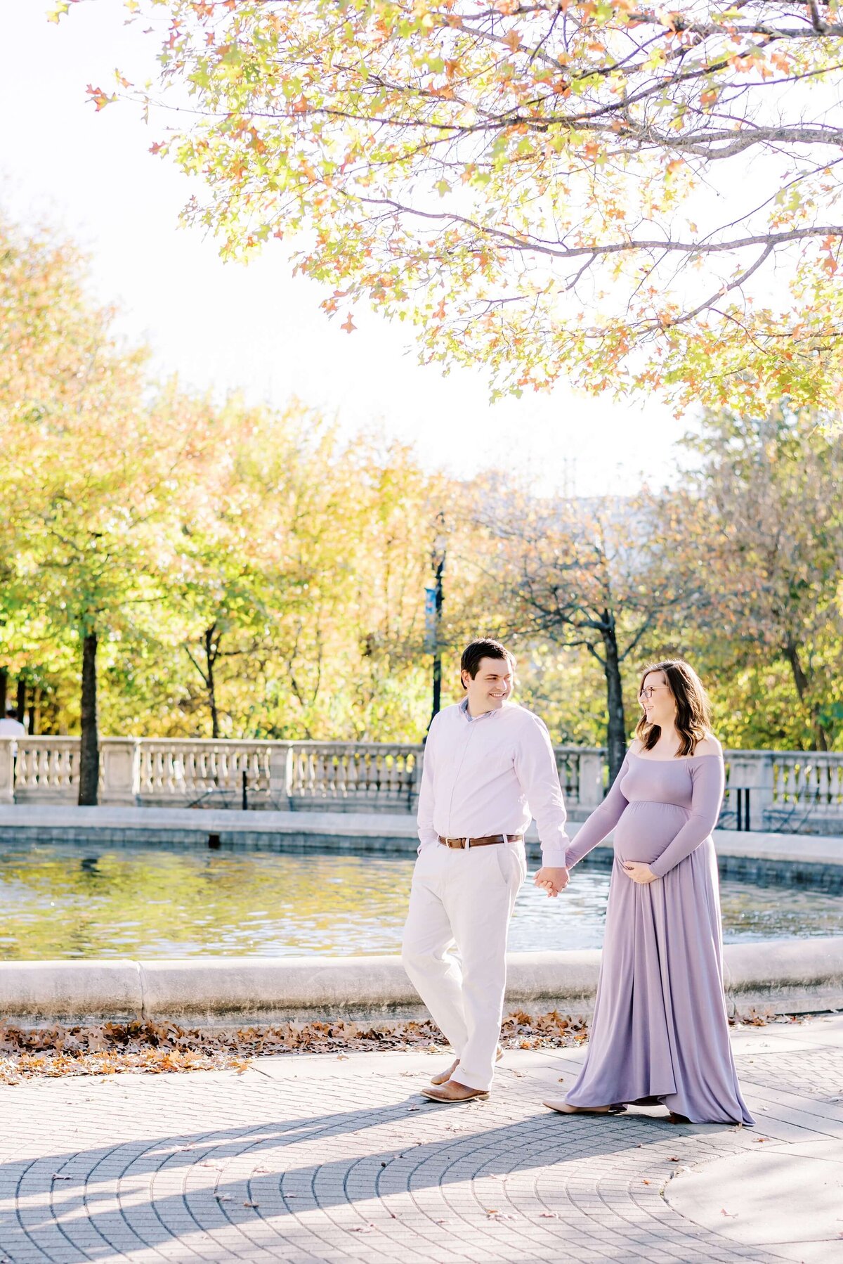 An expecting couple hold hands while taking a stroll through Charlotte's Midtown Park during the fall.