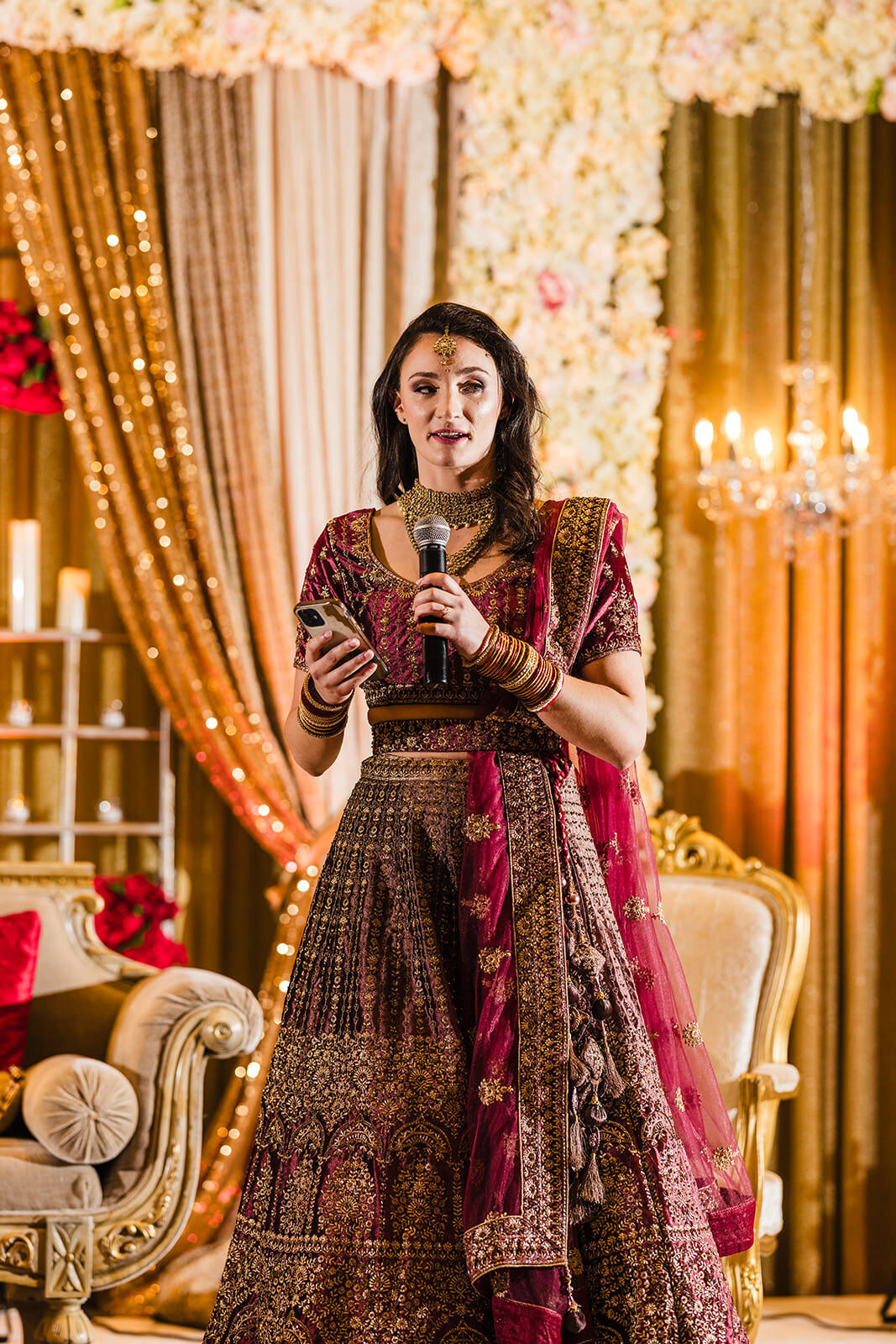 A bride dressed in a traditional burgundy lehenga speaks into a microphone, standing before an ornate wedding stage.