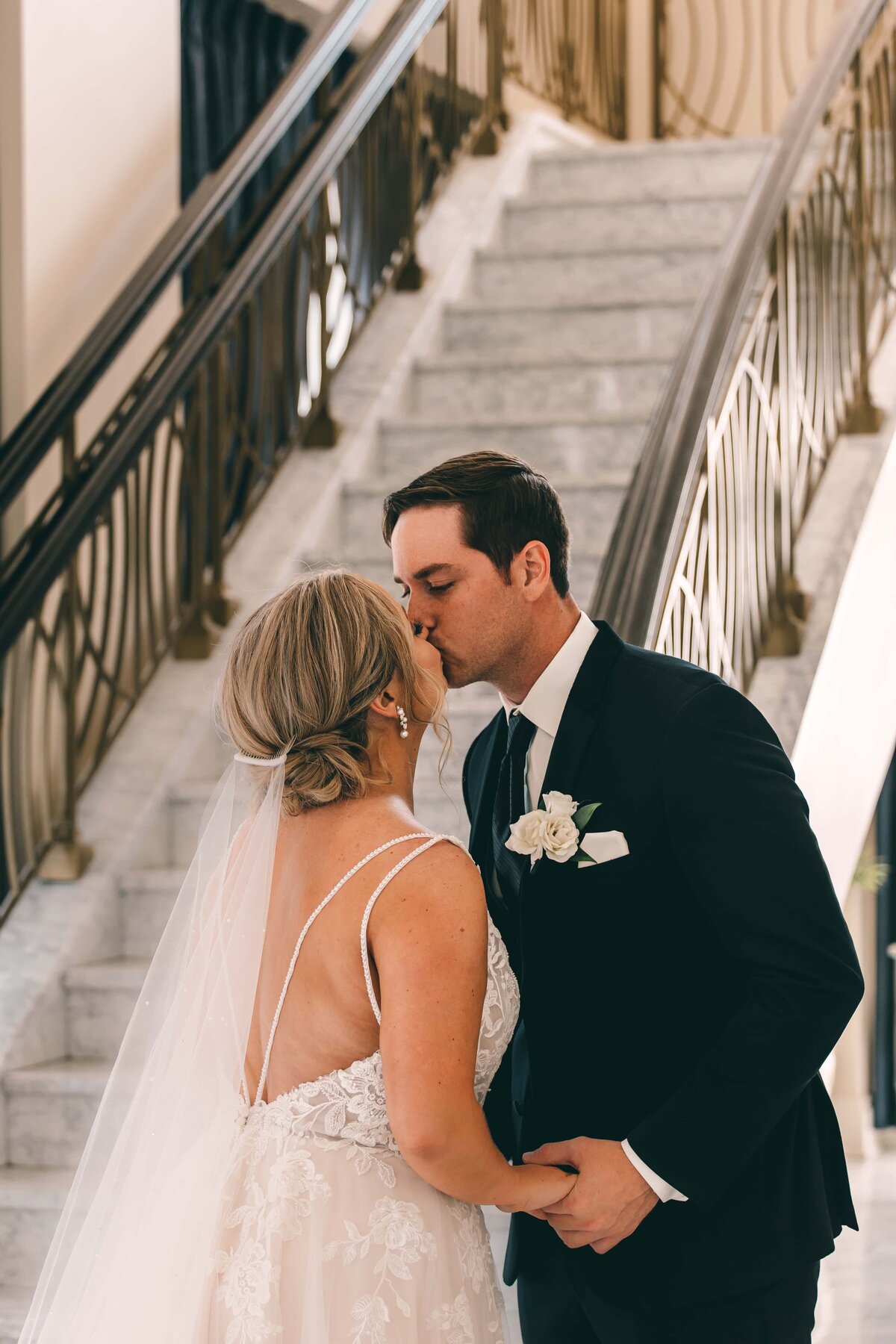 A bride and groom share a kiss on a staircase at their Iowa wedding, with the groom dressed in a black suit and the bride in a white lace gown with a veil.