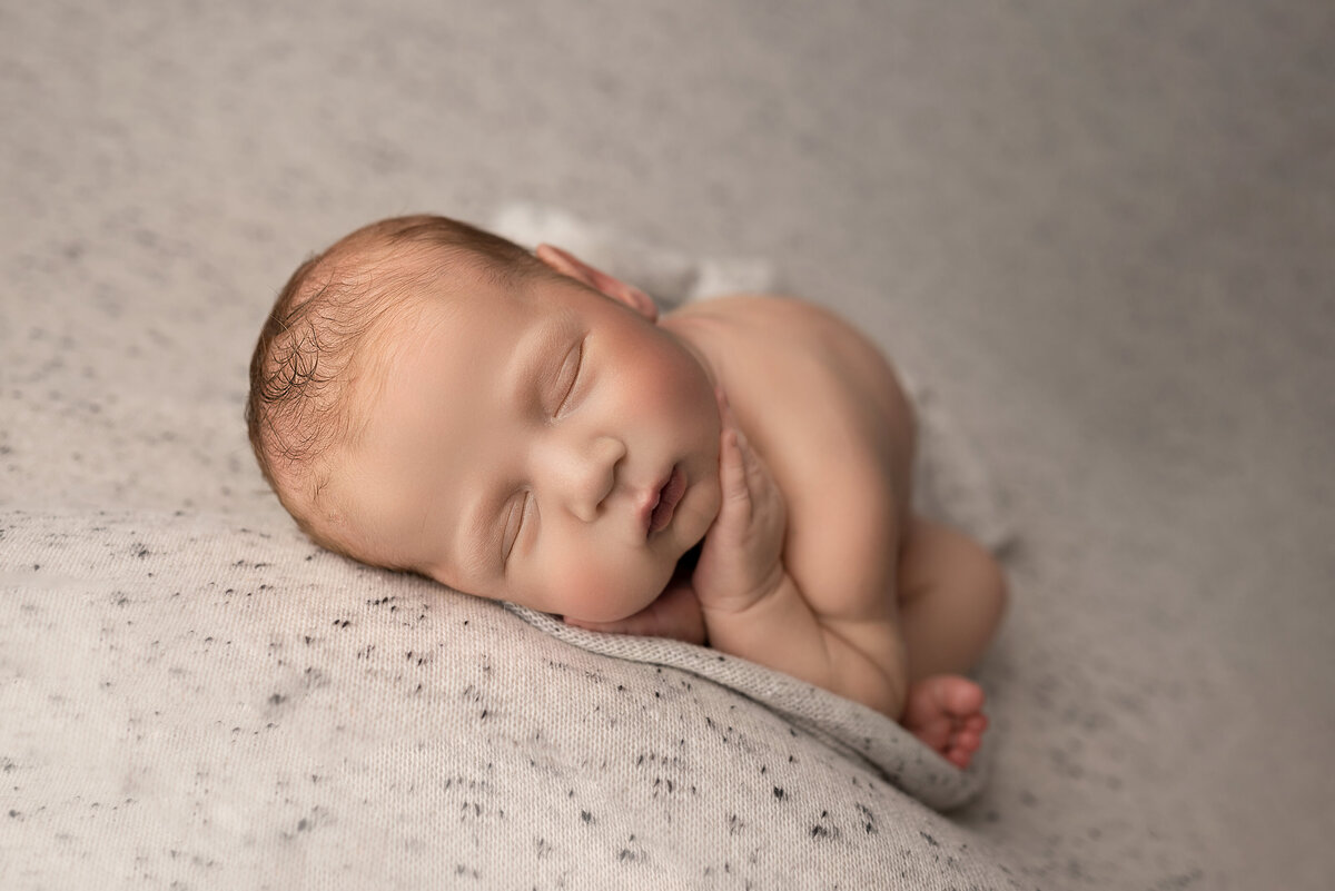 New Jersey's best newborn photographer, Katie Marshall captures an image of baby sleepign on his side with his feet folded underneath him. The baby's hand is resting delicately atop of his cheek.
