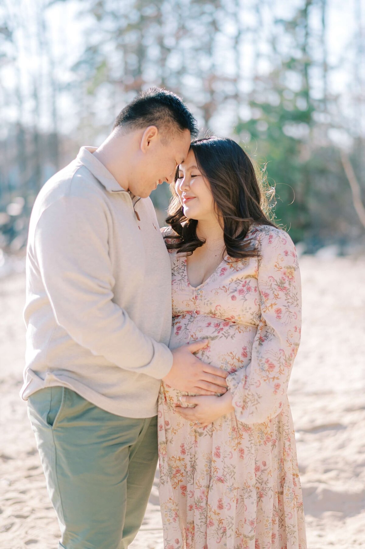 A couple lean their foreheads together as they lightly hold her baby bump while standing on a sandy beach with trees in the background.