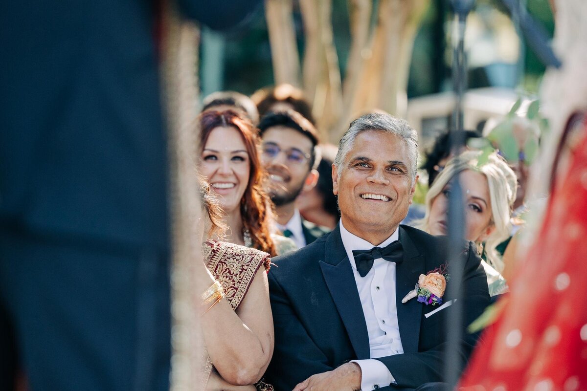 Guests smiling at a wedding ceremony, focused on a middle-aged man in a tuxedo with a boutonniere.