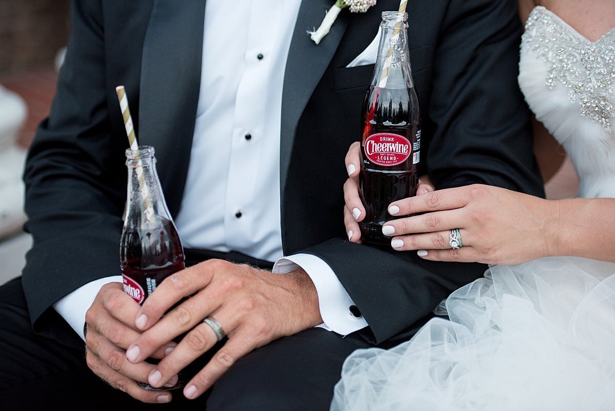The bride and groom show off their wedding rings. The groom, wearing a black tuxedo with a white shirt holds a glass bottle of Cheerwine with a gray and white striped paper straw.  His silver wedding band is on display. Th e bride, wearing a strapless dress with a ruffled organza skirt with  pearls and rhinestones on the bodice holds a glass bottle of Cheerwine with a gray and white paper straw. Her arm is linked through the grooms as she shows off her diamond engagement ring and matching wedding band.