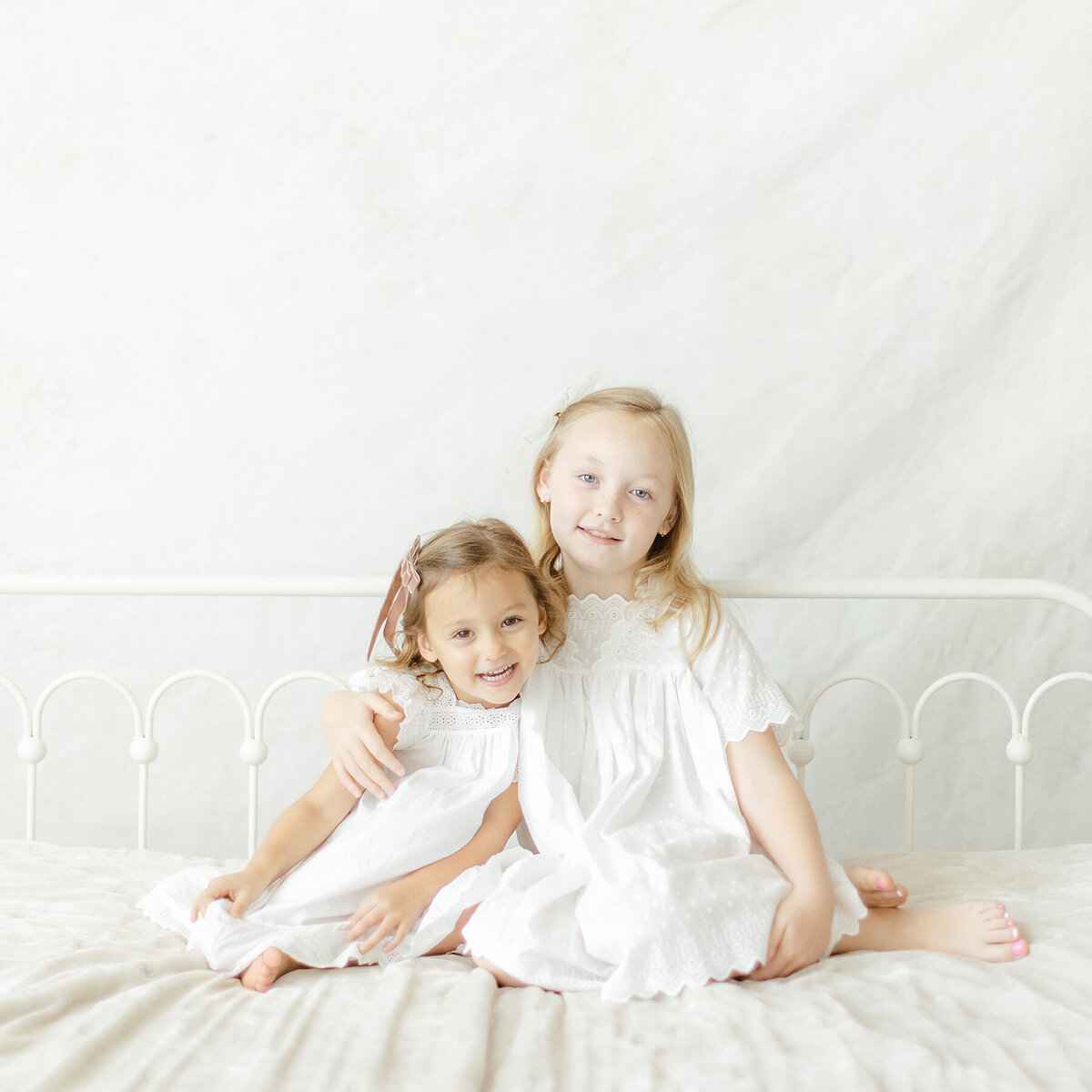 2 little sisters sitting on a bed all snuggled together in a Dallas photography studio for their family portraits.