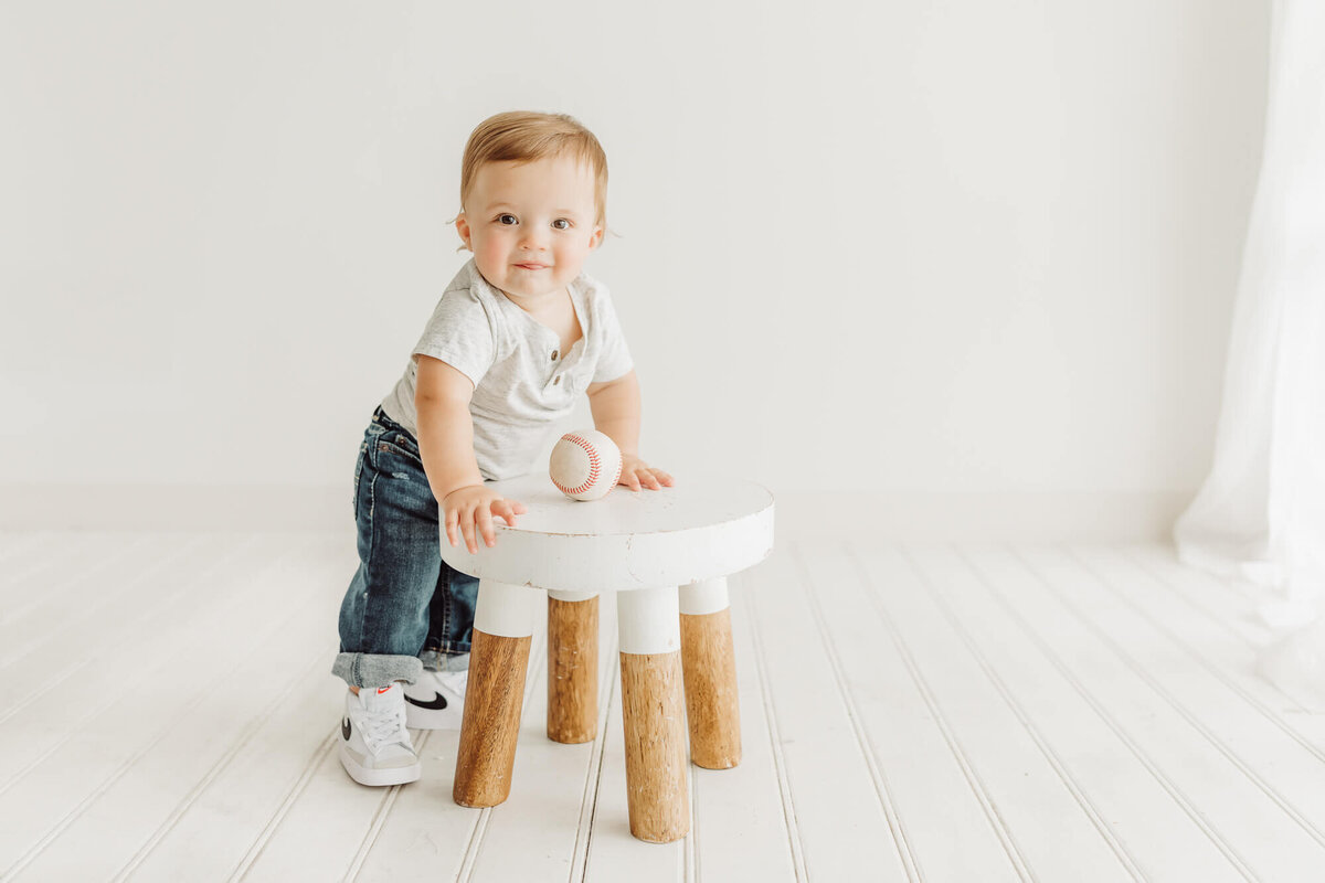 little boy standing against a stool with a baseball for his one year pictures.