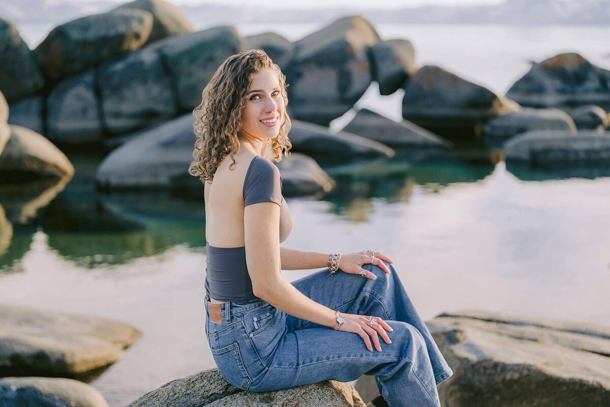 Senior portrait session at Lake Tahoe with grad girl looking at camera sitting on a boulder at water's edge at lake Tahoe. Winter session looking more like summer, no snow