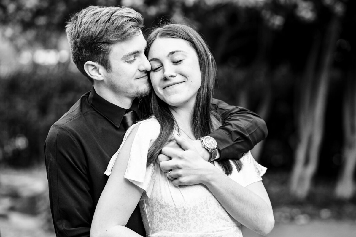 Black-and-white-image-of-engaged-heterosexual-couple-the-man-has-his-arms-around-the-woman-nuzzling-into-her-temple-their-eyes-are-closed-and-both-are-smiling-amid-the-foliage-of-the-McGill-Rose-Garden-in-Charlotte