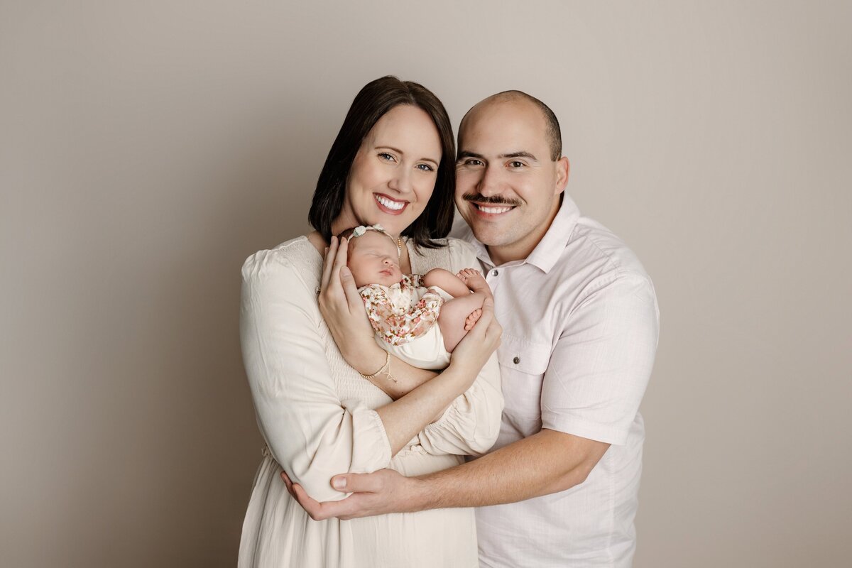 mother holds her newborn daughter in her arms with dad beside her on a cream studio background