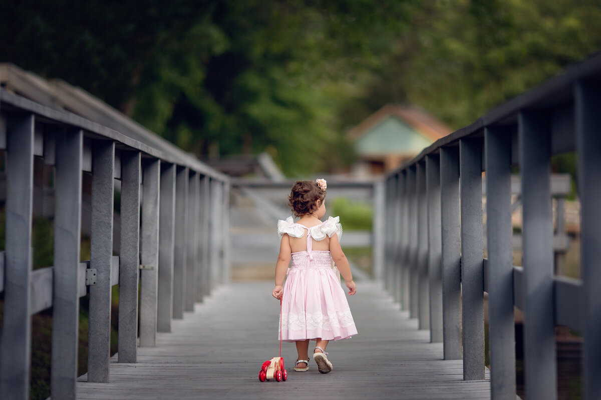 A toddler girl in a pink dress walks her wooden toy across a wooden bridge in a park