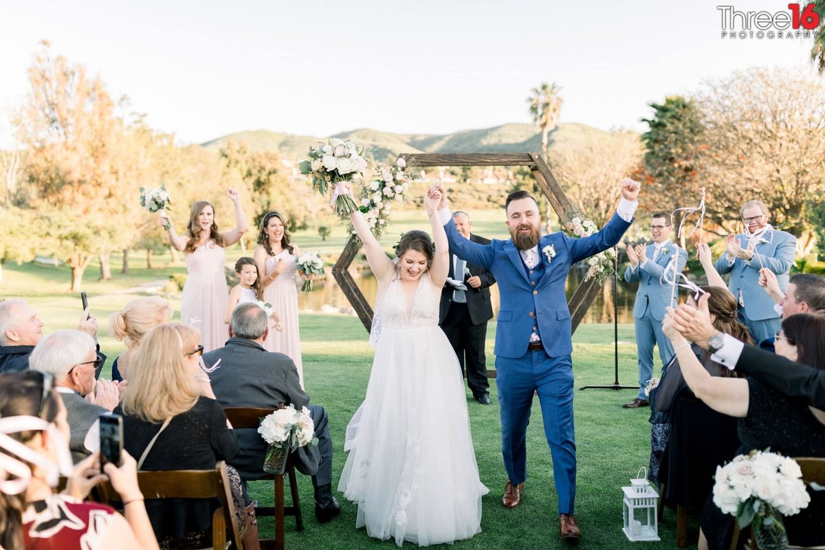 Bride and Groom lift their arms in the air in celebration as they walk down the aisle amongst their guests at outdoor ceremony