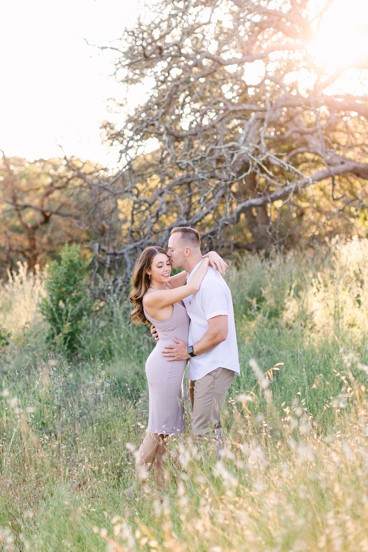 engaged couple hugging in sunny open grass field with trees in background.