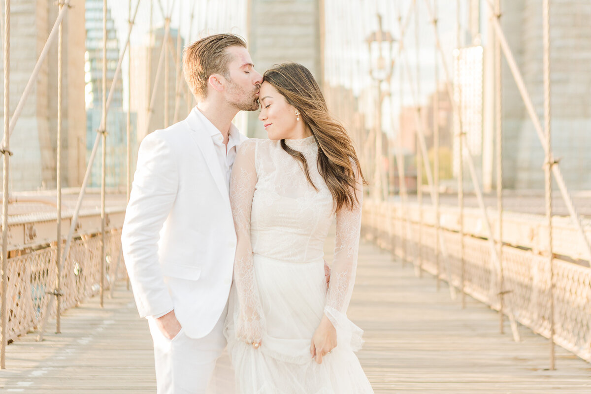 A couple stands on the Brooklyn Bridge in NYC for formal wedding poses. Captured at sunrise, there are no other pedestrians on the bridge. The bride is facing the camera with the groom standing with his chest behind her arm. Her head is turned toward him and he is kissing her on her forehead. Captured by best NYC elopement and wedding photographer Lia Rose Weddings