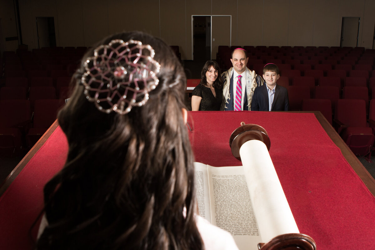 Two parents and brother are seen smiling at their daughter pictured from behind reading from the Torah scroll at Orangetown Jewish Center Rockland bar mitzvah photographer Milestone Images