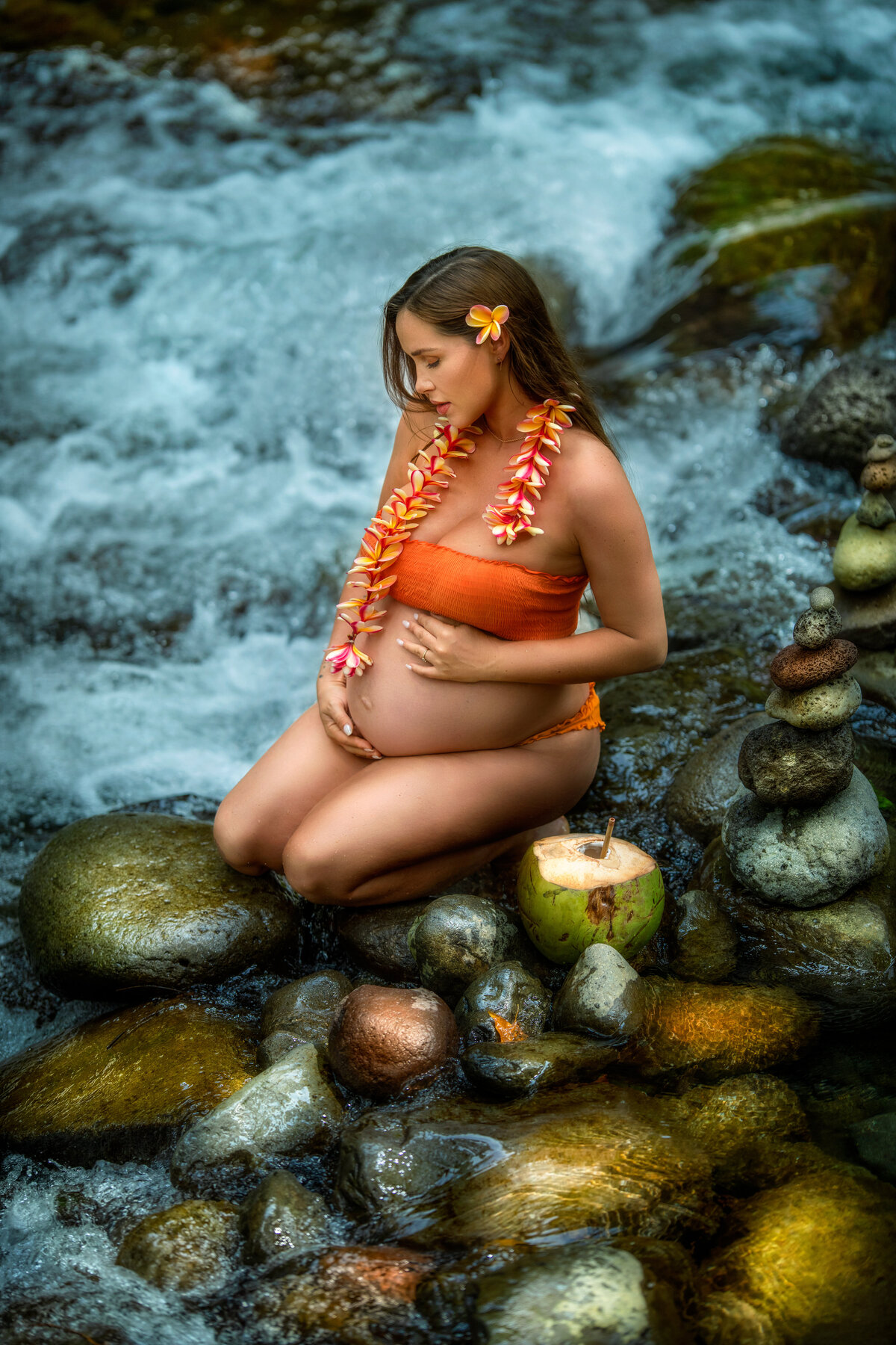 Pregnant  woman kneeling on the rocks in the water by waterfall with coconut and flower necklace in Maui Hawaii