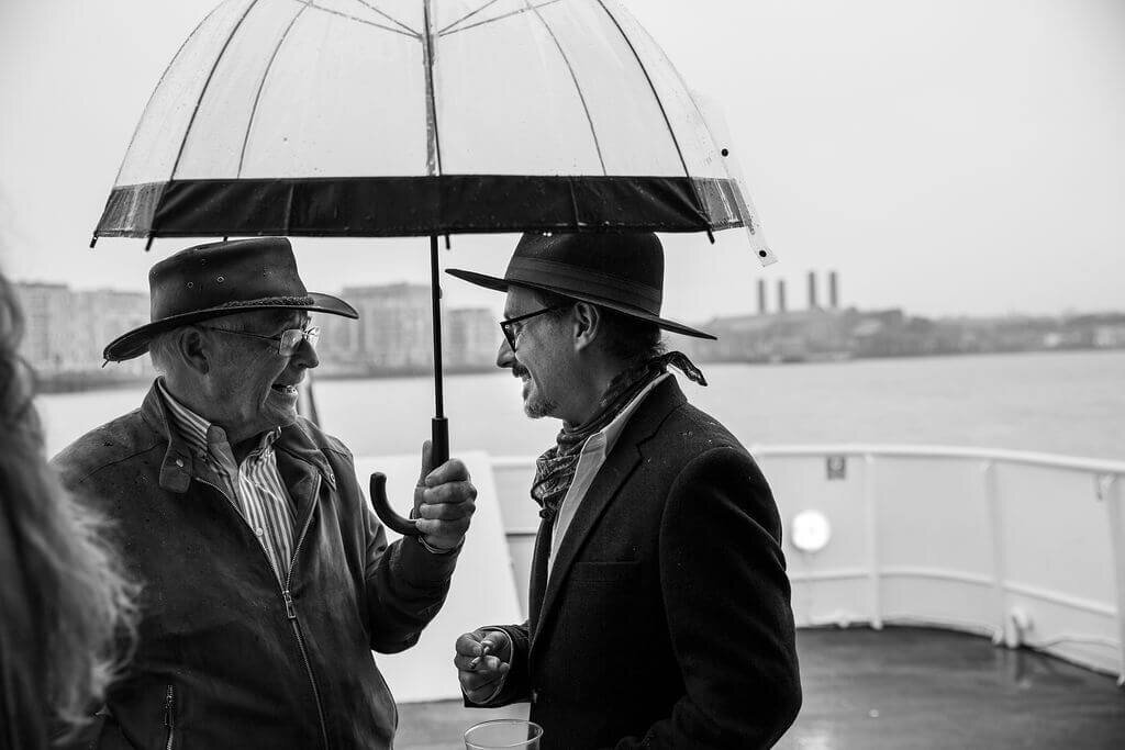 Two men wearing hats hold an umbrella in the rain on the thames
