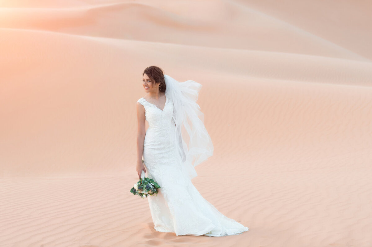 Bride in a lace wedding dress and a floral bouquet standing amidst Arabian desert dunes, elopement photoshoot organized by Lovely & Planned