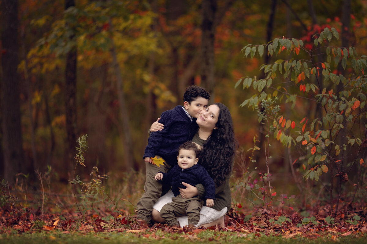 A happy mother kneels in a park in fall with her two toddler sons in matching blue sweaters