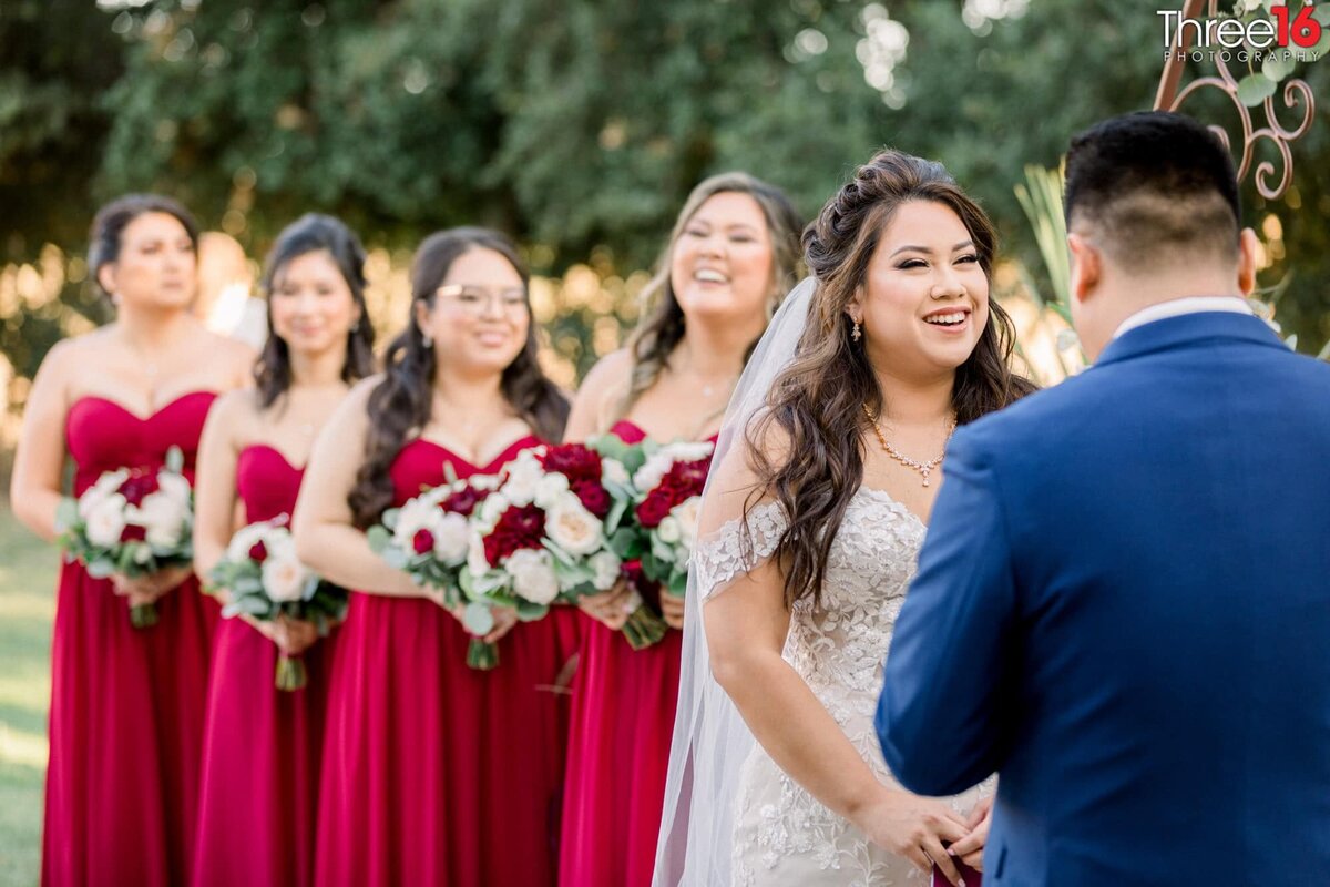 Bride smiles at her Groom as he reads his vows to her while bridesmaids look on