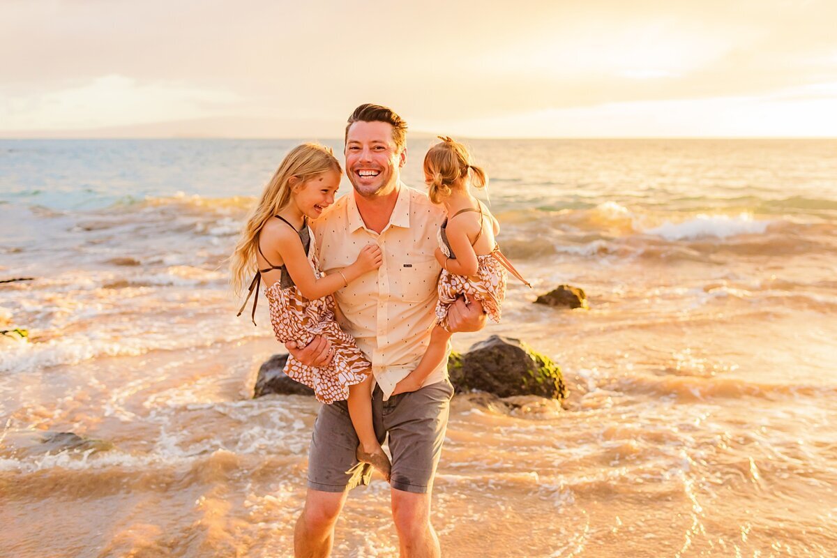 Dad holds daughters on his hips at the beach while smiling at sunset with Love + Water
