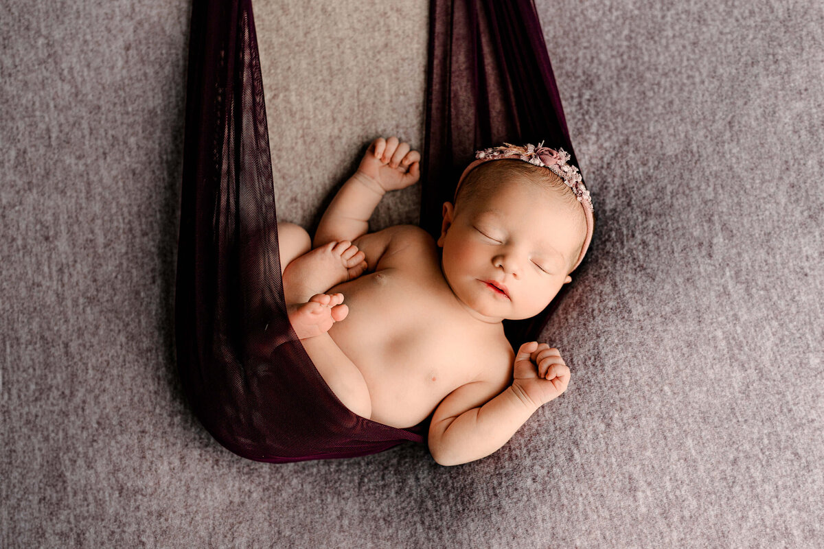 newborn baby girl posed in a  purple sheer cloth swing wearing a purple headband
