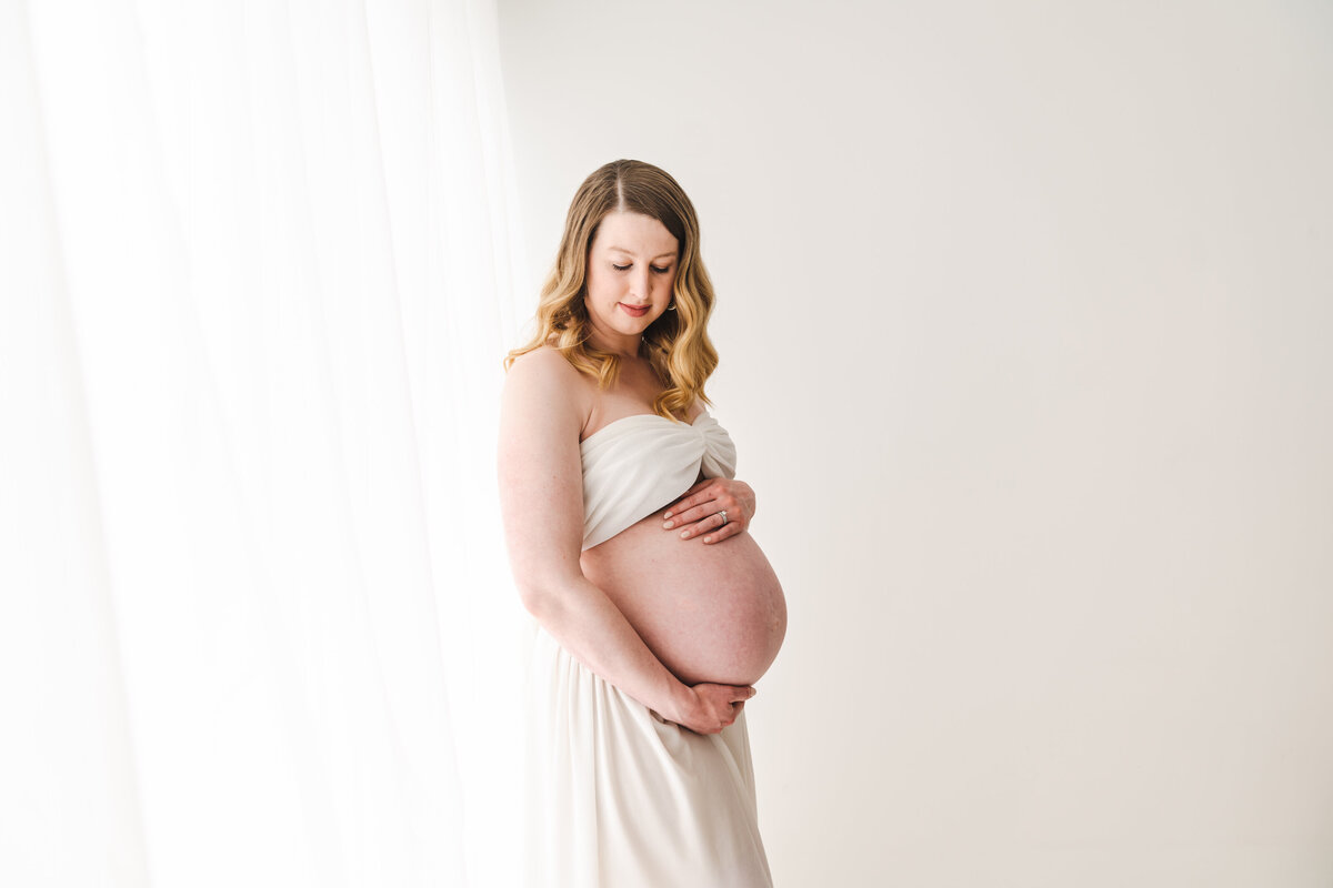 A radiant expecting mother in a white dress tenderly cradling her baby bump against a bright, airy backdrop. Taken by Fig and Olive Photography, a Minneapolis Maternity Photographer.