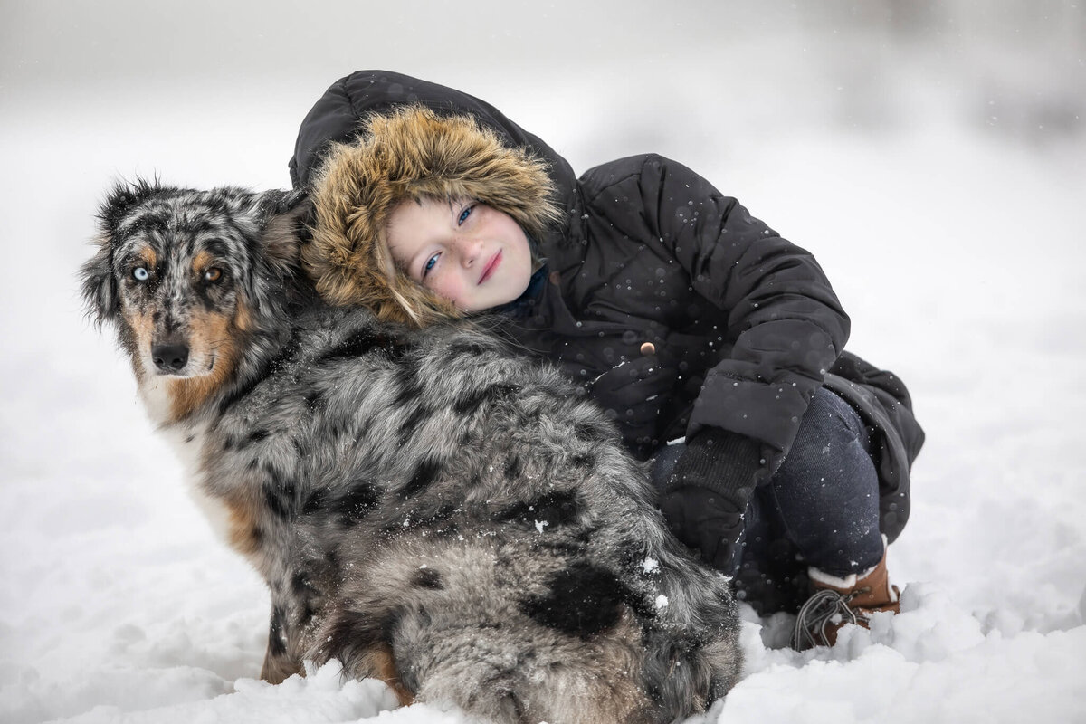 Girl is wearing a black hooded, fur lined coat  snuggled with her Australian Shepherd while  being photographed in snow and both are looking at the camera