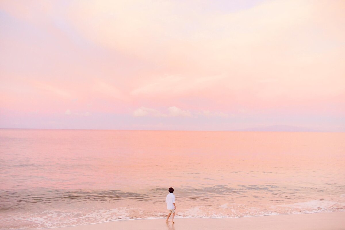 Artistic family portrait of young boy walking toward the ocean at sunrise with pink pastel skies above him