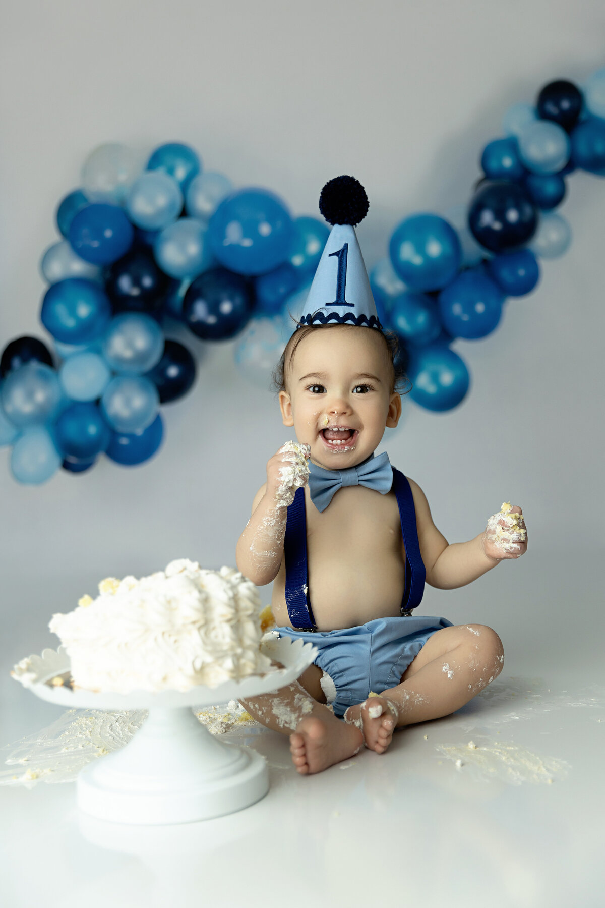 A happy toddler sits on a New Jersey Cake Smash Photographer studio floor covered in cake wearing blue suspenders and bowtie