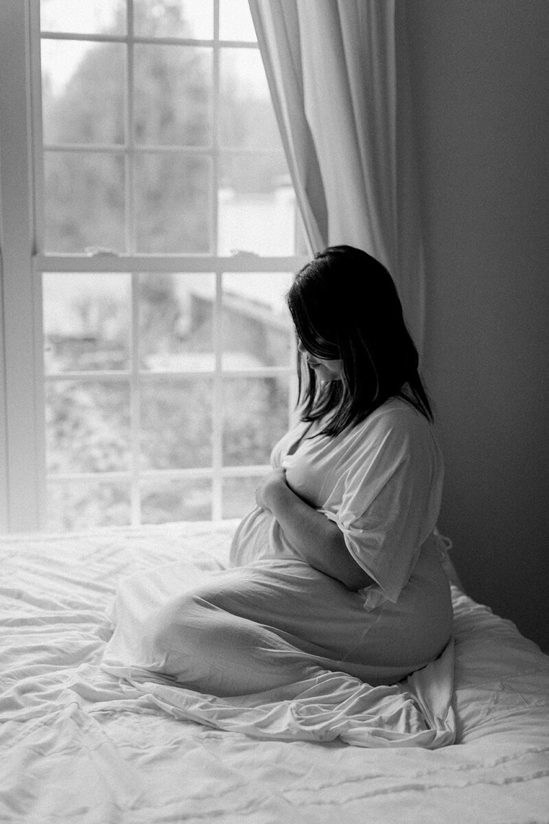 black and white photo of a pregnant asian mom sitting on a bed by the window