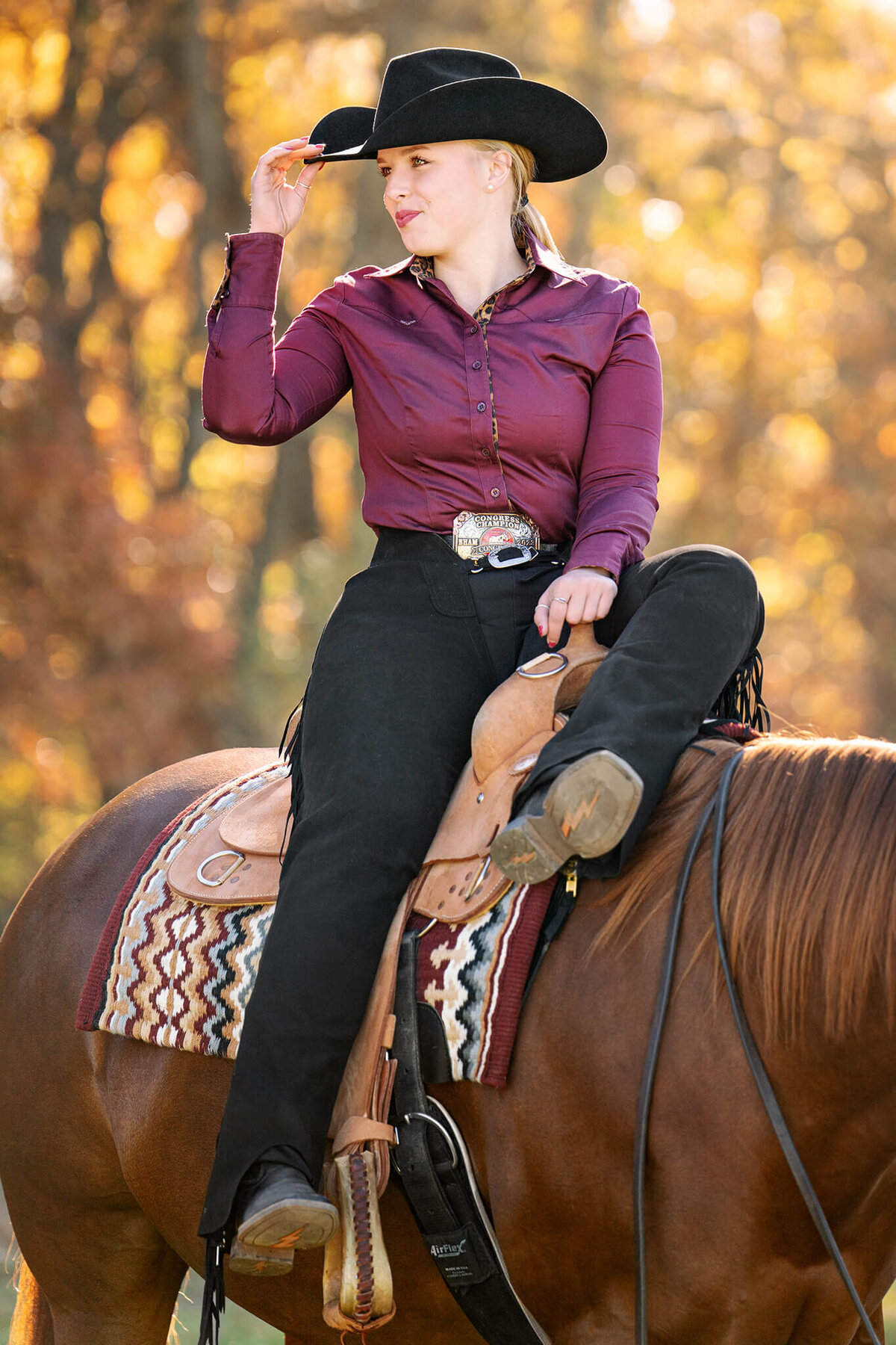 blonde equestrian wearing a purple shirt and black chaps sitting sideways on her chestnut paint horse grasping the bill of her cowboy hat. behind them is beautiful yellow and orange leaves.