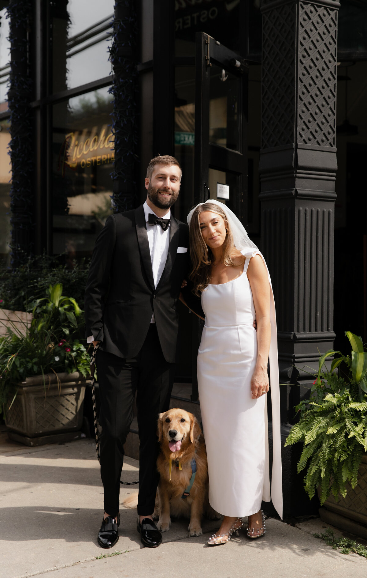 Bride in simple, modern, white dress and veil stands next to groom in tuxedo outside of Elske Restaurant with their dog at wedding ceremony.