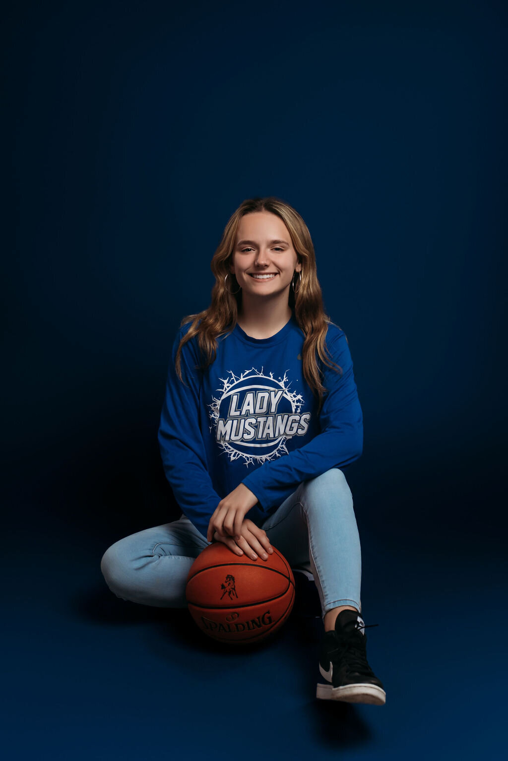 A teenage girl in a sports warmup sits on the floor and holds a basketball in front of a blue solid backdrop for her senior graduation photoshoot, captured by a fine art studio photographer in Bemidji, Minnesota