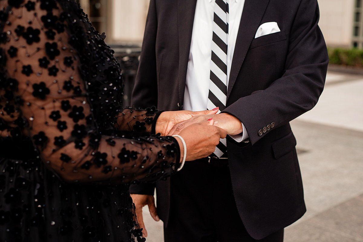 The bride puts the groom's wedding ring on his finger. The groom is wearing a black suit with a white shirt and a black and white striped tie. He has a white pocket square in his lapel pocket. The bride is wearing a long sleeved sheer white dress with small black flowers.