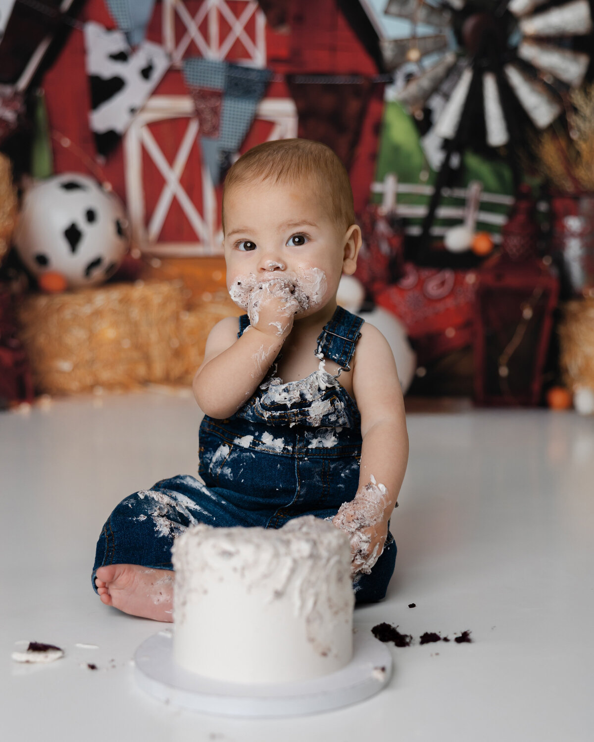 Baby boy enjoying a messy cake smash in front of a red barn backdrop with rustic farm props. Photograph by Yucaipa cake smash photographer Kaitlyn Dawn Photography.