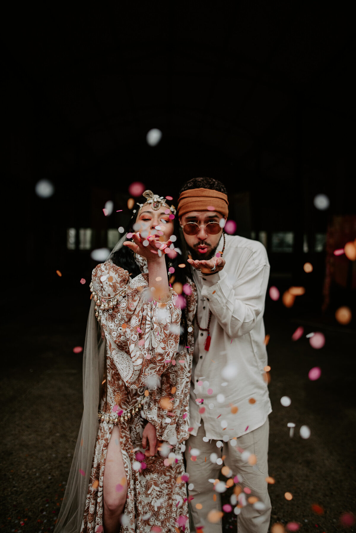 A bride and groom blow confetti into the camera at Happy Valley Norfolk. The wedding was styled with a 70s vibe.