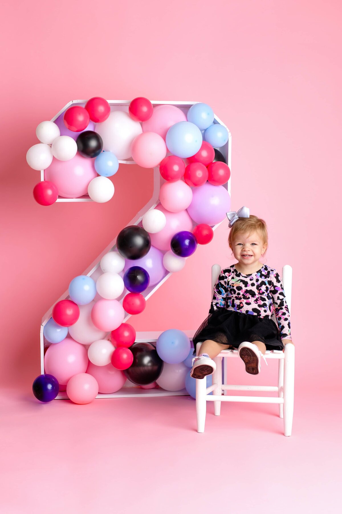 Little girl sitting in a multicolored dress with a black bottom with a large number two prop sitting beside her filled with balloons for her second birthday celebration