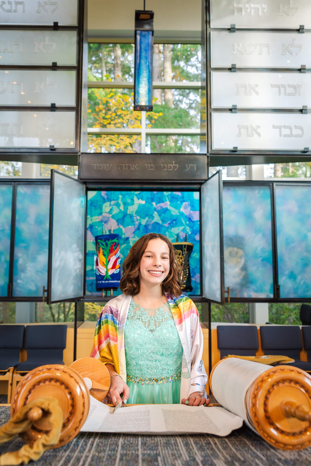 A happy teenage girl in a green dress and rainbow tallit stands at the bimah with the large torah