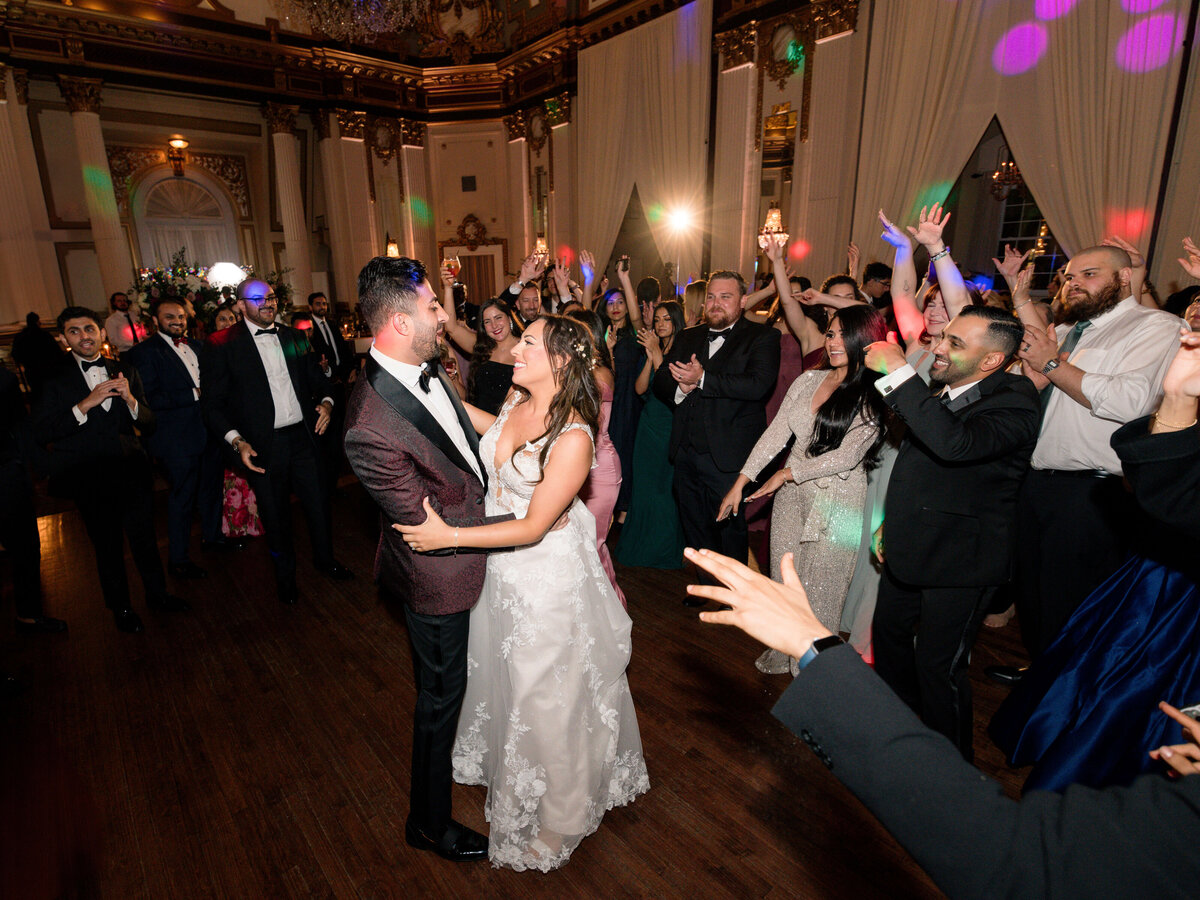 A bride and groom dance in the center of a ballroom, surrounded by cheering guests. The groom wears a dark suit, and the bride is in a white gown. The room is elegantly decorated, and colorful lights add to the celebratory atmosphere.