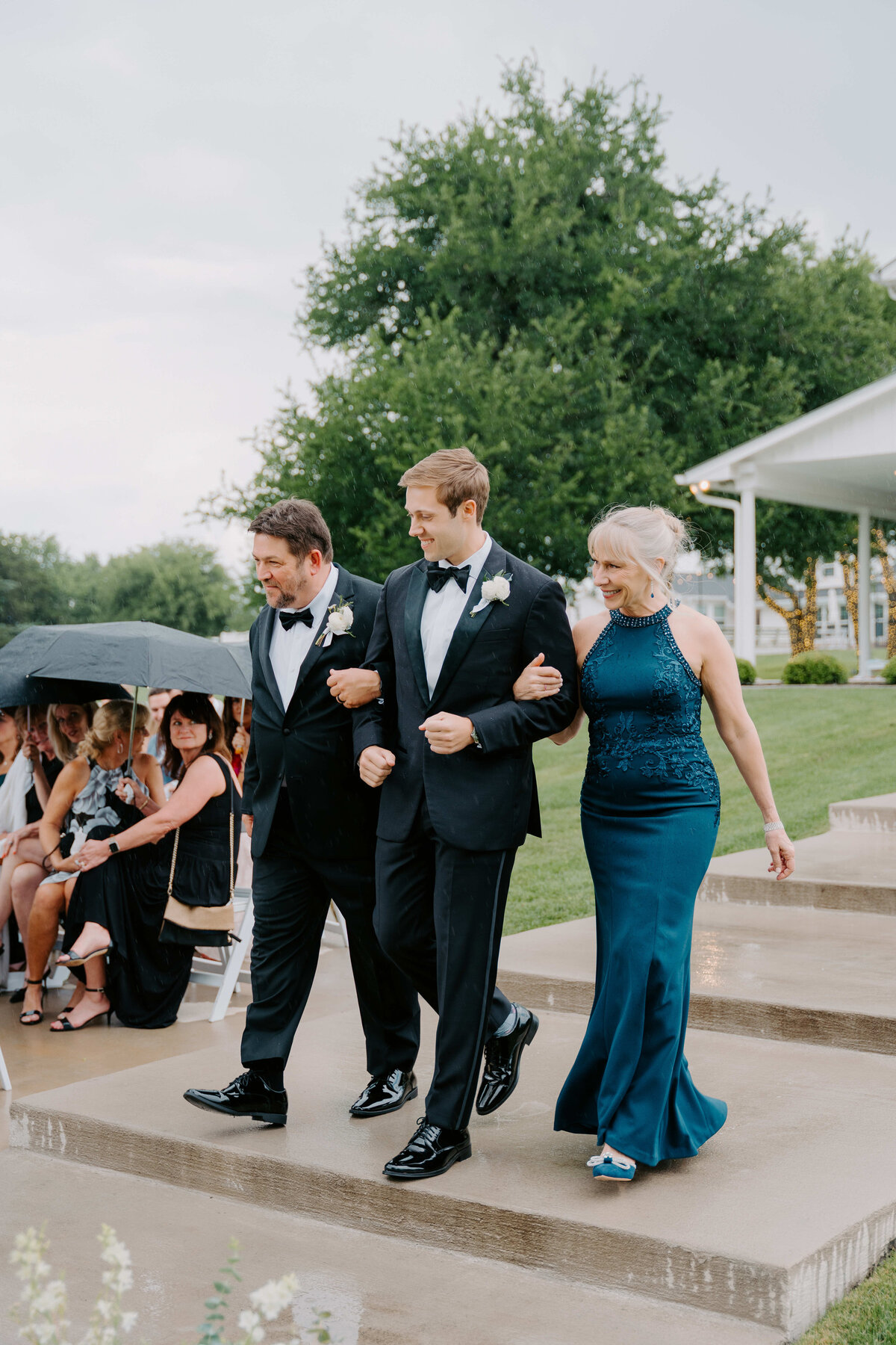 A groom in a black tuxedo walks arm-in-arm with two elegantly dressed individuals in blue formal wear. People under umbrellas watch on, conveying a joyful, rainy wedding scene.
