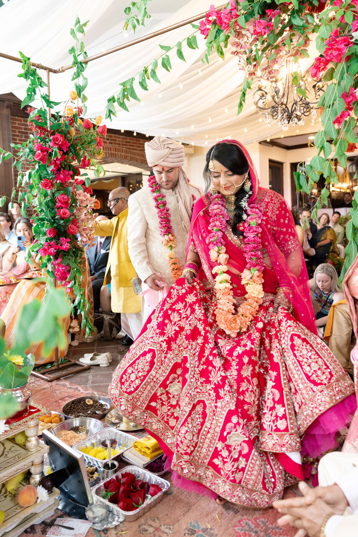 A couple in traditional Indian attire participate in a wedding ceremony. They are adorned with floral garlands under a canopy decorated with greenery and flowers, surrounded by guests. Ritual items are placed in front of them.