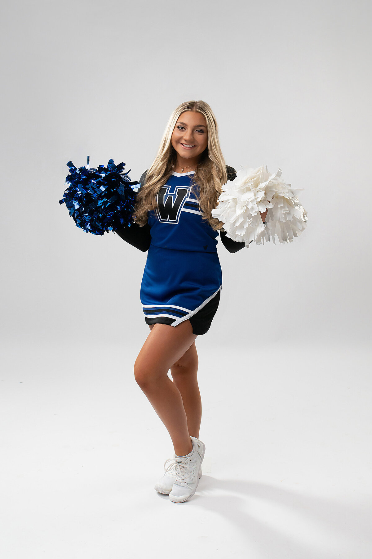 A senior student from Waukesha West High School stands in our photo studio wearing her cheerleading uniform and holding poms.