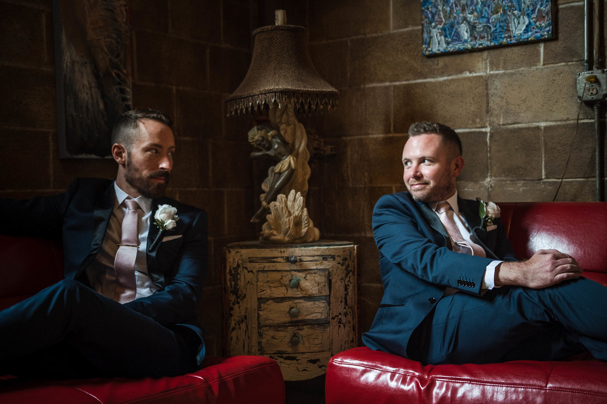 Grooms sit on couches and smile at each other on wedding day.